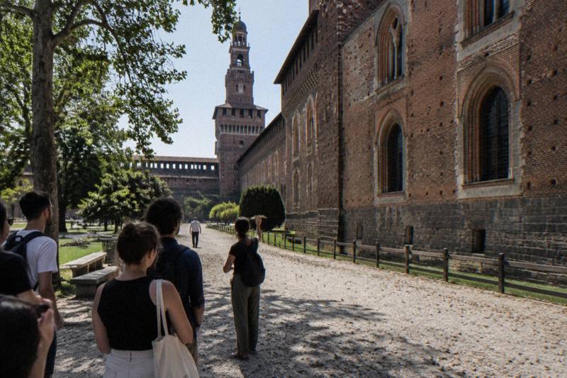 Midday at Castello Sforzesco in Milan, Italy, a free walking tour paused beneath the fortress's rust-red brick ramparts and arched stone windows, harsh sun overhead, gravel path scattered with plane tree seeds, a distant figure from behind.