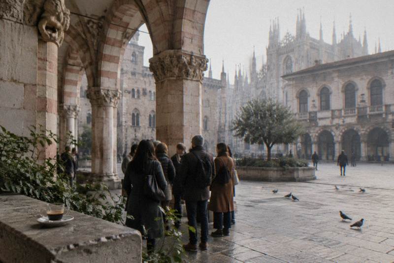A coffee culture walk gathers under the pink-and-white marble arcades of the Loggia degli Osii in Piazza Mercanti, Milan, Italy on a hazy morning, medieval stone carvings overhead, a half-finished espresso left on a ledge, pigeons on the paving.