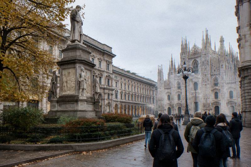 A travelers' choice free tour starts by the Manzoni monument on Piazza San Fedele in Milan, Italy on a crisp autumn morning, granite statue and classical facades in soft grey light, fallen leaves along the stone gutters, distant backs of walkers.