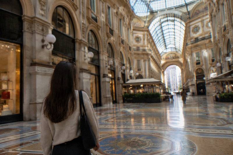 Afternoon light pours through the glass vault of the Galleria Vittorio Emanuele II in Milan, Italy, a walking tour standing in the mosaic-paved octagon, worn marble floor gleaming, one distant shopper out of focus near a cafe doorway.